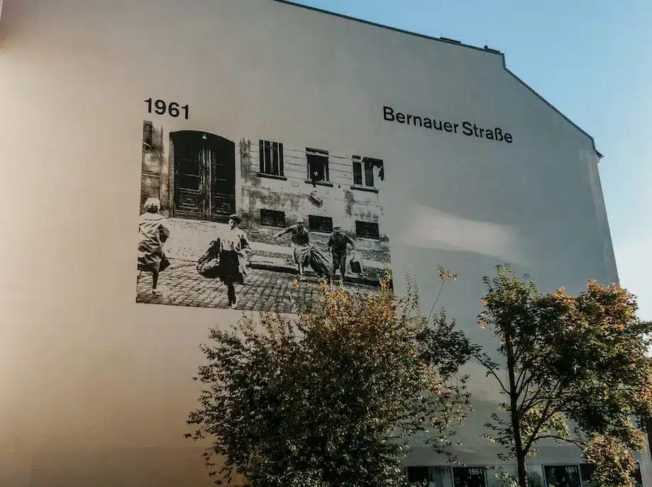Exposição de fotografia gigante, reproduzida em um muro na Bernauer Strasse, onde se vê quatro pessoas atravessando a rua em Berlim em 1961