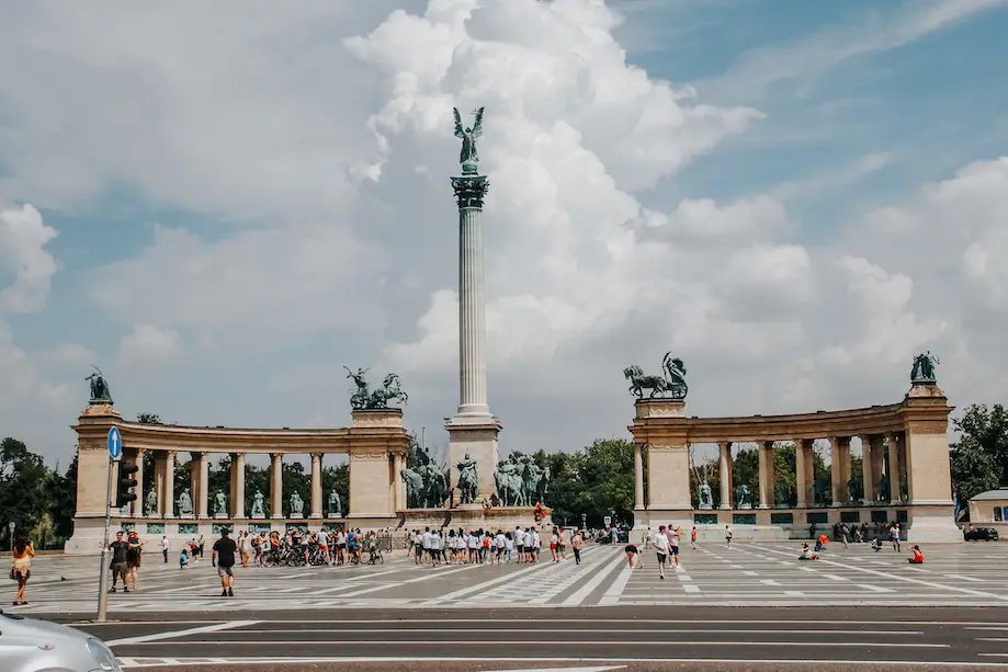 Foto das esculturas na Praça dos Heróis, em Budapeste, com um obelisco no centro e pilastras formando um arco ao redor dele