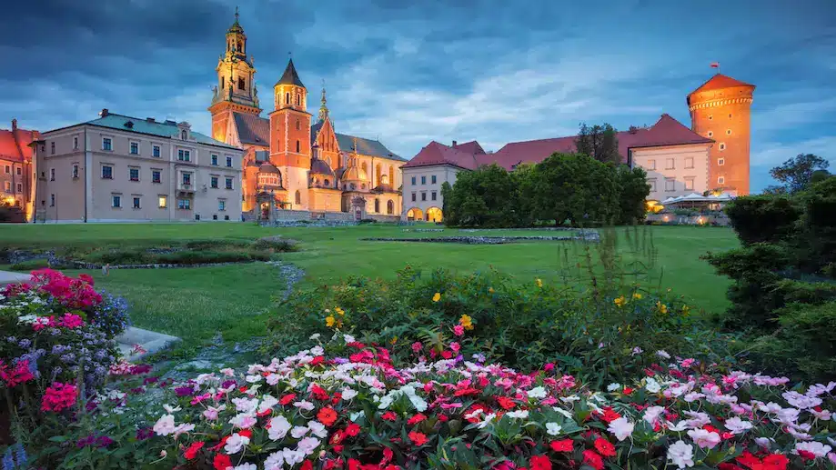 Foto noturna doda Cidade Velha de Cracóvia, com um jardim na frente e as construções iluminadas ao fundo. Esse é o melhor lugar para se hospedar em Cracóvia.