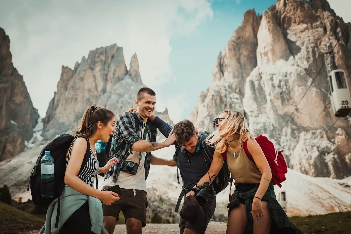 Worldpackers - Voluntariado Internacional: grupo de quatro jovens com mochilas e câmeras se abraçando e rindo em frente a uma montanha