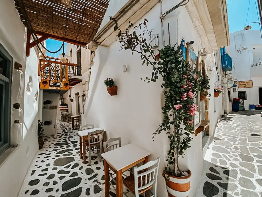 Imagem de um beco no centro histórico de Naxos Town, com casinhas brancas, ruas estreitas, cadeira de restaurante para fora e uma roseira na parede